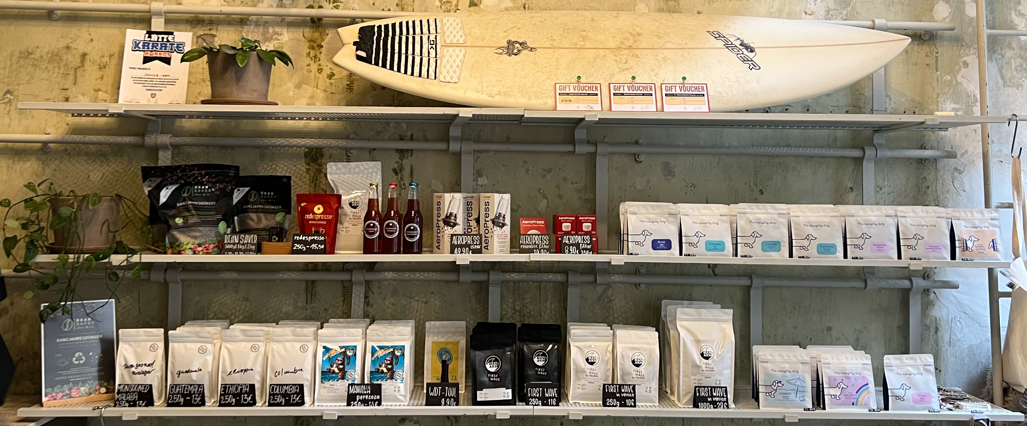 Ein Regal im MANANA café in Wien 1180 Währing mit verschiedenen Kaffeeprodukten, darunter Kaffeebohnen, AeroPress-Sets, Geschenkgutscheine, Red Espresso und Kombucha, dekoriert mit einem Surfbrett und Pflanzen.